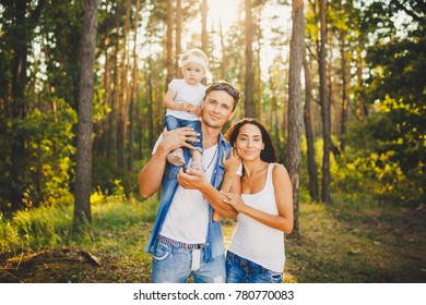 Stylish Young Family Of Mom, Dad And Daughter One Year Old Blonde Sitting Near Father On Shoulders, Outdoors Outside The City In A Park Amid Trees In Summer. Wear Jeans Clothes. Family Photo Session