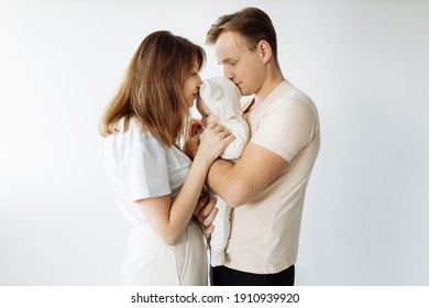 Stylish Young Family, Photographed With A Little Beautiful Baby. Dad Holds The Baby In His Arms, Mom Kisses It Gently. The Family Expresses Love And Awe To Each Other. Family And Parenting Concept.