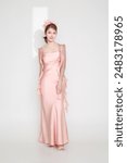 Stylish young fashionable Asian woman in a stunning pink dress, trendy makeup with light shining through a slit on her, posing on an isolated white background.