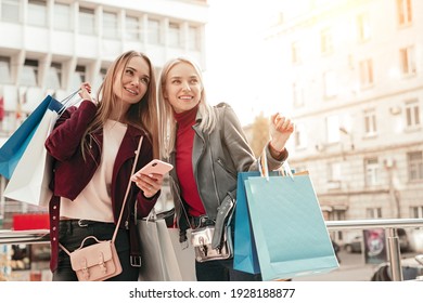 Stylish young female friends in trendy clothing with shopping bags using mobile phone while checking information about sales during shopping time together in city - Powered by Shutterstock