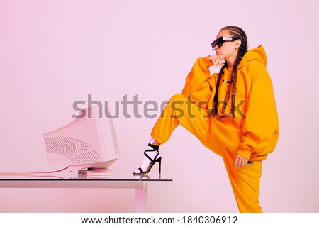 Stylish young female office worker in sunglasses with old computer monitor and foot on desk. Colored light