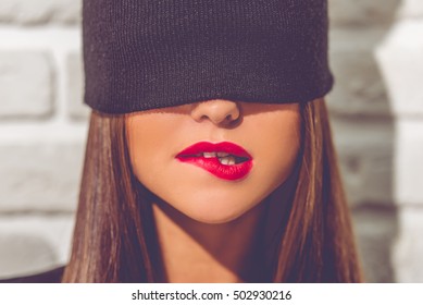 Stylish young girl with a black cap on her eyes is biting her red lips, on brick wall background