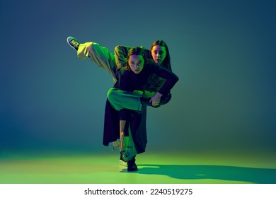 Stylish young girls dancing casual clothes dancing experimental style dance over gradient blue-green background at dance hall in neon light. Youth culture, contemporary dance, fashion, action.