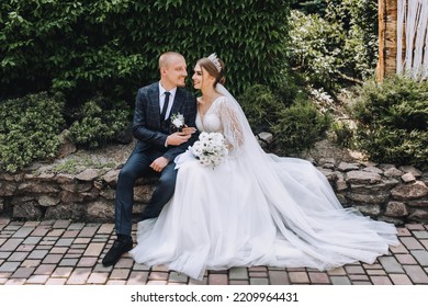 A stylish, young groom and a beautiful bride in a long white dress with a bouquet in their hands, a diadem on their heads, sit gently hugging in a park in nature. Wedding photography of the newlyweds.