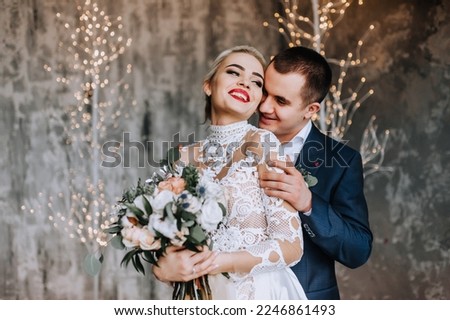 Stylish young groom in a blue suit hugs a beautiful smiling blonde bride in a white lace dress in a studio, interior against the background of bokeh lamps. Wedding photography, portrait.
