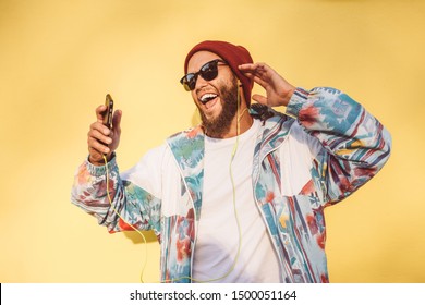 Stylish young hipster man with beard in red hat and a retro jacket of 90s on yellow background dancing and singing while he listens to the music in his smartphone. Relax. love live - Shutterstock ID 1500051164