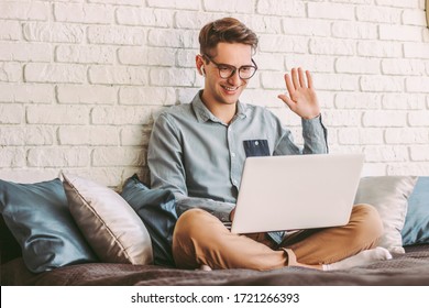 Stylish Young Hipster Man Student In Glasses Waving Hand To Laptop While Talking Online Video Call And Sitting On Couch. Happy Businessman Freelancer Having Web Meeting, Conference. Work At Home
