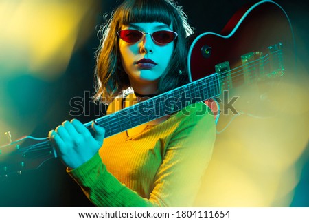 Stylish young hipster woman with curly hair holding red guitar on shoulder in neon lights. 90s style concept.