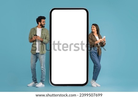 Stylish young indian man and woman using phones and looking at huge smartphone with white empty screen, checking newest mobile app, isolated on blue studio background, mockup, blank space