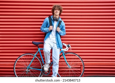 Stylish young male in casual clothes keeping hands on backpack straps while leaning on red metal wall and bike and looking at camera