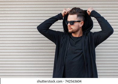 Stylish Young Man With A Beard In Sunglasses In A Black Hoody Dresses A Hood Near A Metal Wall