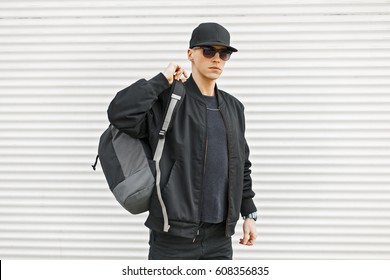Stylish young man in black modern clothes with a backpack near a white wall
