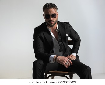 Stylish Young Man In Black Tuxedo With Open Collar Shirt Holding Elbow On Knee  And Posing On Grey Background In Studio