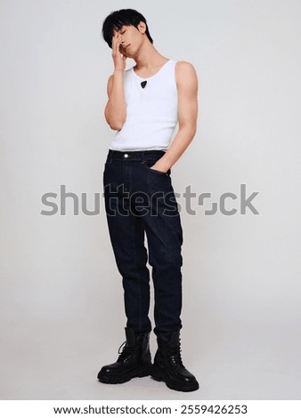 Stylish Young Man in Casual White Tank Top, Dark Denim Jeans, and Black Combat Boots Striking a Thoughtful Pose Against a Neutral Background.