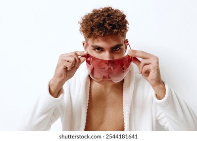 Stylish young man with curly hair wearing a red transparent face mask, posing confidently in a white robe against a clean, bright background. Fashion and self-expression concept. - Powered by Shutterstock