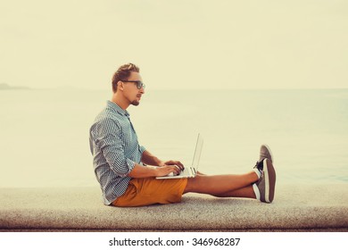 Stylish Young Man In Glasses With A Beard Shirt And Shorts And Sneakers Working On Laptop While Sitting On The Beach On The Pier, Sending Mail, Mounts, Video Editing And Photography