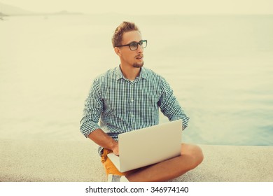 Stylish Young Man In Glasses With A Beard Shirt And Shorts And Sneakers Working On Laptop While Sitting On The Beach On The Pier, Sending Mail, Mounts, Video Editing And Photography