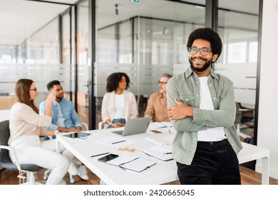 A stylish young man with glasses and a green shirt confidently stands with arms crossed in bright office, his colleagues discuss work in background. Blend of professionalism and a relaxed atmosphere - Powered by Shutterstock