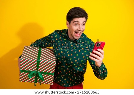 A stylish young man in a green patterned shirt holds a striped present and smiles at a red smartphone against a bold yellow backdrop, conveying holiday shopping celebration and festive lifestyle