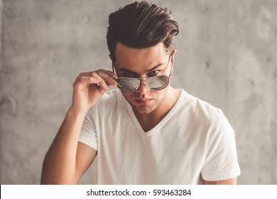 Stylish Young Man Is Looking At Camera From Under Sun Glasses, On Concrete Wall Background