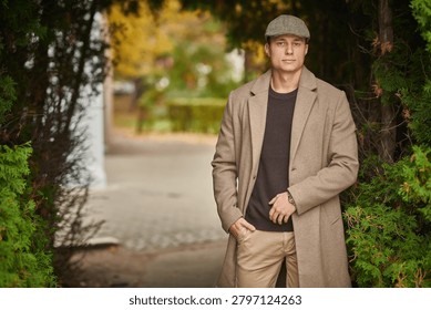 Stylish young man posing between evergreen trees in a beige coat and flat cap. Sophisticated autumn fashion with natural framing and copy space.