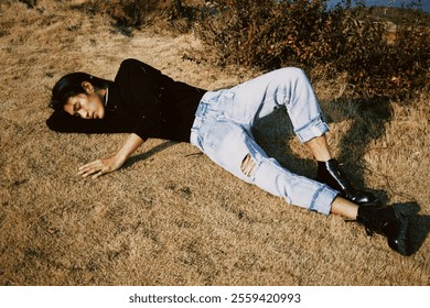 Stylish Young Man Relaxing Outdoors in Casual Ripped Jeans and Black Boots, Enjoying a Sunny Day on a Dry Grassy Field. - Powered by Shutterstock