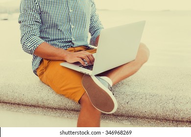 Stylish Young Man In A Shirt And Shorts And Sneakers Working On Laptop While Sitting On The Beach On The Pier, Sending Mail, Mounts, Video Editing And Photography