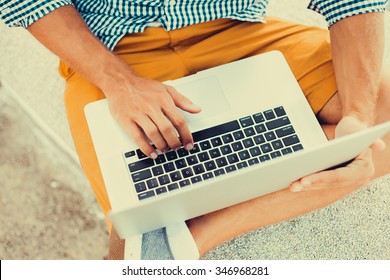 Stylish Young Man In A Shirt And Shorts And Sneakers Working On Laptop While Sitting On The Beach On The Pier, Sending Mail, Mounts, Video Editing And Photography