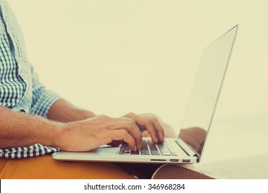 Stylish Young Man In A Shirt And Shorts And Sneakers Working On Laptop While Sitting On The Beach On The Pier, Sending Mail, Mounts, Video Editing And Photography