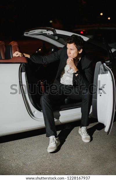 Stylish young man\
sitting posing in his white cabriolet. Nightlife. Businessman in\
suit in luxury car