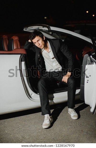 Stylish young man\
sitting posing in his white cabriolet. Nightlife. Businessman in\
suit in luxury car
