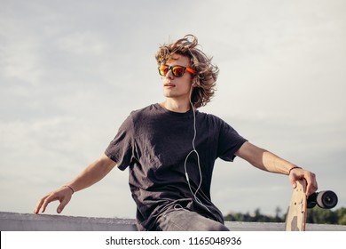 stylish young man with skateboard listening music with headphones , near concrete wall
