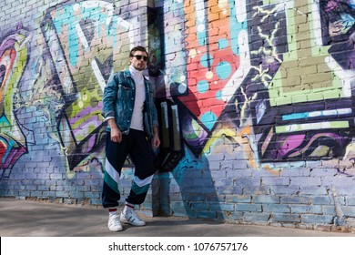 Stylish Young Man In Vintage Denim Jacket And Track Pants Leaning On Brick Wall With Graffiti