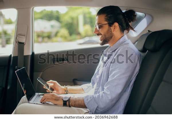 Stylish young\
middle-eastern guy independent contractor working while going\
somewhere by car, long-haired arab man sitting at auto back seat,\
using laptop and mobile phone, copy\
space
