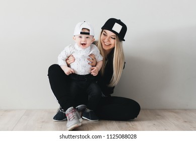 Stylish Young Mom In Cap Sitting And Having Fun With Her Cute One Year Old Baby Isolated On White Background. Sports Family