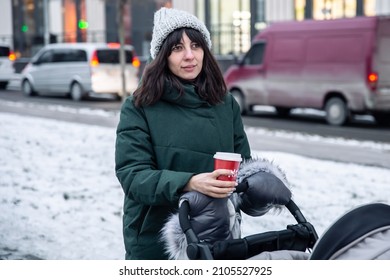 Stylish Young Mom With A Cup Of Coffee On A Walk With Her Baby In A Baby Carriage In Winter.