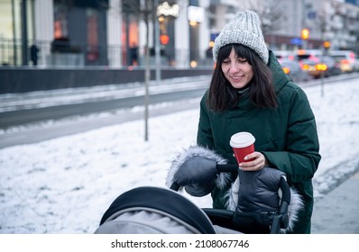 Stylish Young Mom With A Cup Of Coffee On A Walk With A Baby Carriage In Winter.