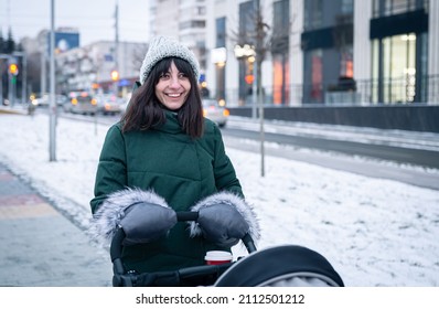 Stylish Young Mom With A Cup Of Coffee On A Walk With A Baby Carriage In Winter.