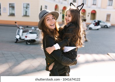 Stylish young mom in retro felt hat carrying her cheerful curly brunette daughter across the street. Portrait of excited girl sitting on mom's hands and looking at her with tenderness - Powered by Shutterstock