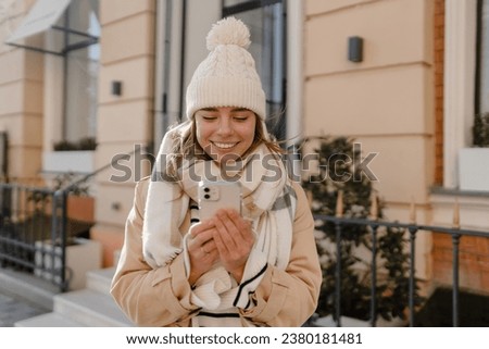 stylish young pretty woman walking in winter street wearing beige coat, knitted hat, scarf, smiling happy cold season fashion trend, using smart phone