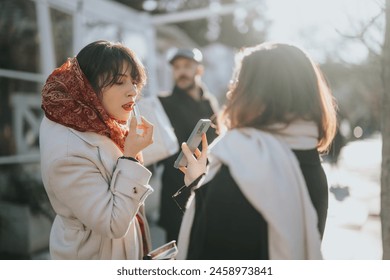 A stylish young woman applies lipstick outdoors with her smart phone in hand as friends converse in the city. A snapshot of urban life and casual gatherings. - Powered by Shutterstock