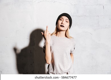 Stylish young woman with black lipstick standing and showing middle finger over white background 