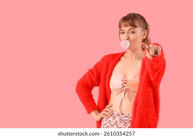 Stylish young woman with chewing gum pointing at viewer on pink background