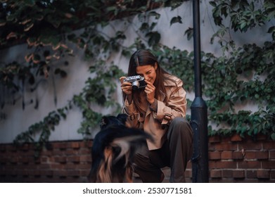 Stylish young woman crouches happily in the city, taking a photo of her dog with a vintage camera among autumn leaves and a brick wall - Powered by Shutterstock