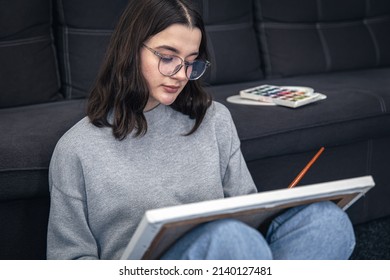 Stylish Young Woman In Glasses, Teenager, Draws A Picture On Canvas While Sitting Near The Sofa In The Interior Of The House.