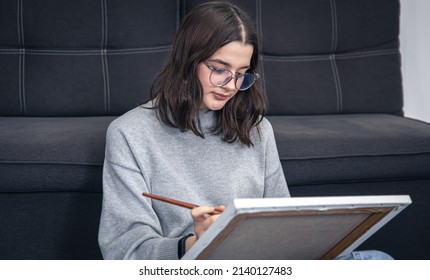 Stylish Young Woman In Glasses, Teenager, Draws A Picture On Canvas While Sitting Near The Sofa In The Interior Of The House.