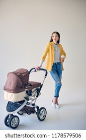 Stylish Young Woman Holding Disposable Coffee Cup While Standing With Baby Stroller And Looking At Camera On Grey