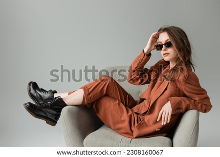 stylish young woman with long hair wearing terracotta suit with blazer, pants and black boots posing in trendy sunglasses while sitting in armchair on grey background