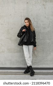 Stylish Young Woman Model In Trendy Rock Outfit With Fashion Leather Jacket, Jeans And Black Leather Shoes With Leather Bag Stands Near A Gray Wall