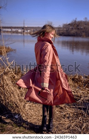 stylish young woman in a pink trench dances on the bank of a spring river                      